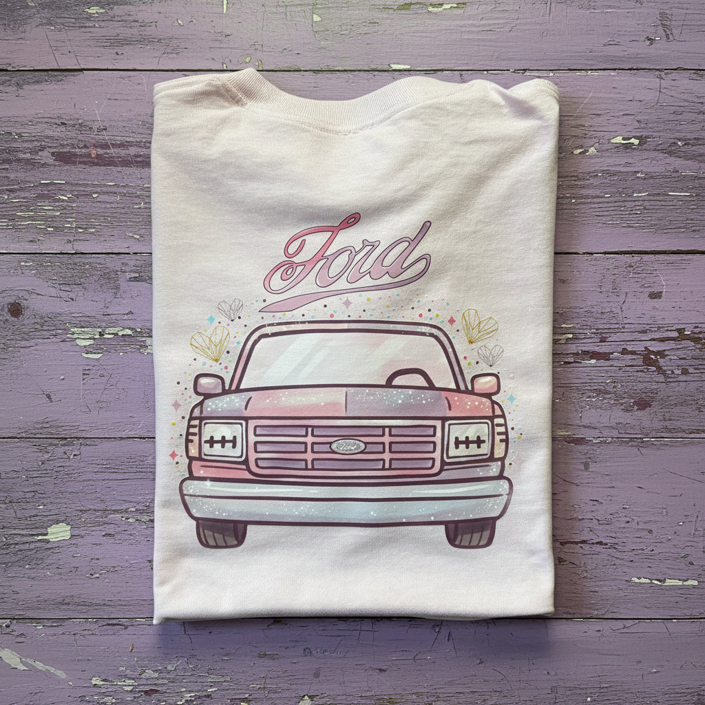 A folded light pink t-shirt featuring a graphic of a vintage Ford pickup truck with a split pink and purple color scheme, and the word 'Ford' written above it, laid flat on a rustic brown wooden background.

