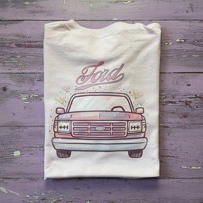 A folded light pink t-shirt featuring a graphic of a vintage Ford pickup truck with a split pink and purple color scheme, and the word 'Ford' written above it, laid flat on a rustic brown wooden background.
