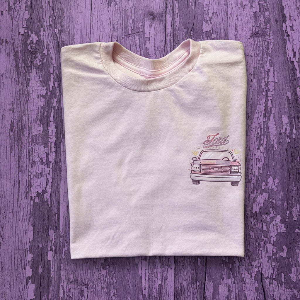 A folded white t-shirt featuring a graphic of a vintage Ford pickup truck with a split pink and purplecolor scheme, and the word 'Ford' written above it, laid flat on a rustic brown wooden background.

