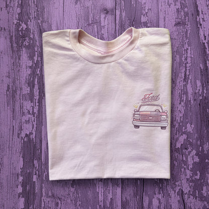 A folded white t-shirt featuring a graphic of a vintage Ford pickup truck with a split pink and purplecolor scheme, and the word 'Ford' written above it, laid flat on a rustic brown wooden background.
